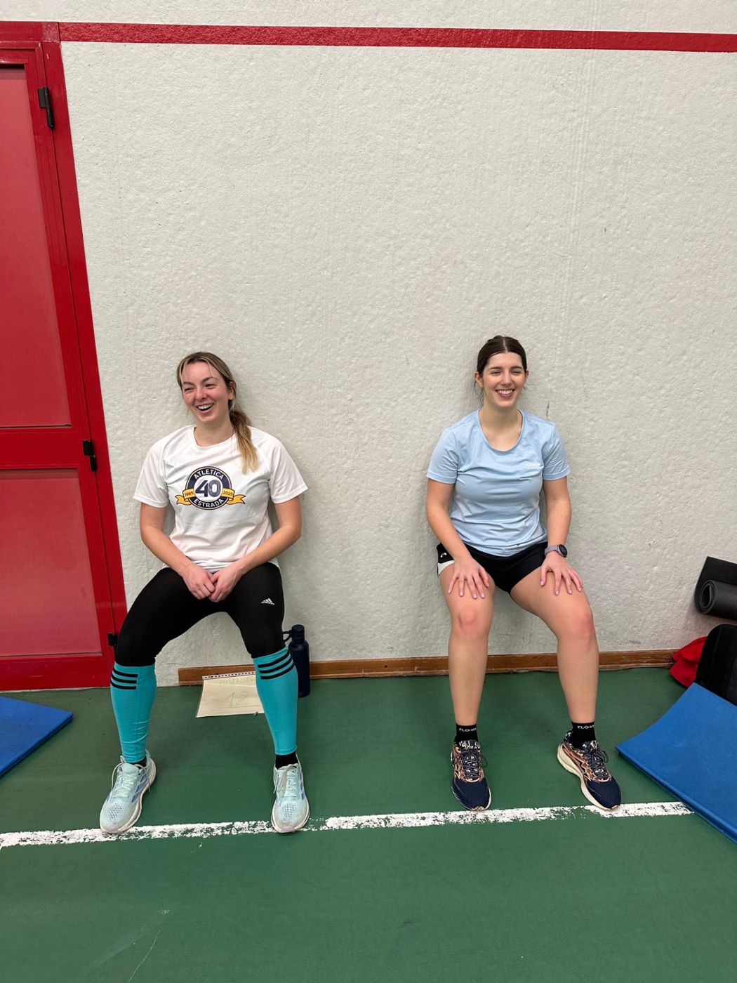 Due atlete eseguono l'esercizio della sedia al muro sorridendo